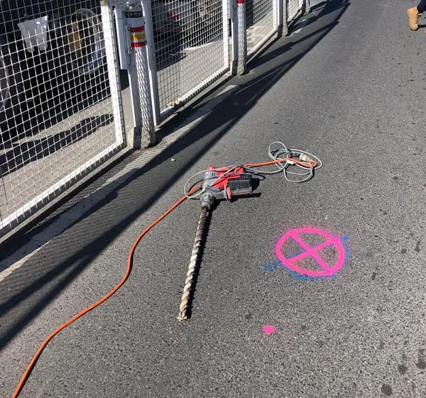A pink cross sprayed on asphalt marking a service-clear bore hole location, with a drill rod beside it