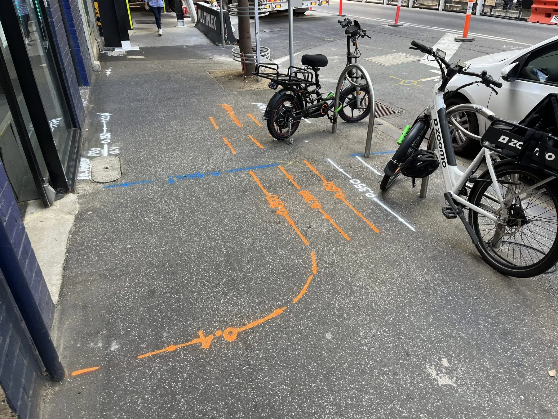 Orange locate lines with painted depths curving across a city footpath, with a blue line and a white line beside parked e-bikes