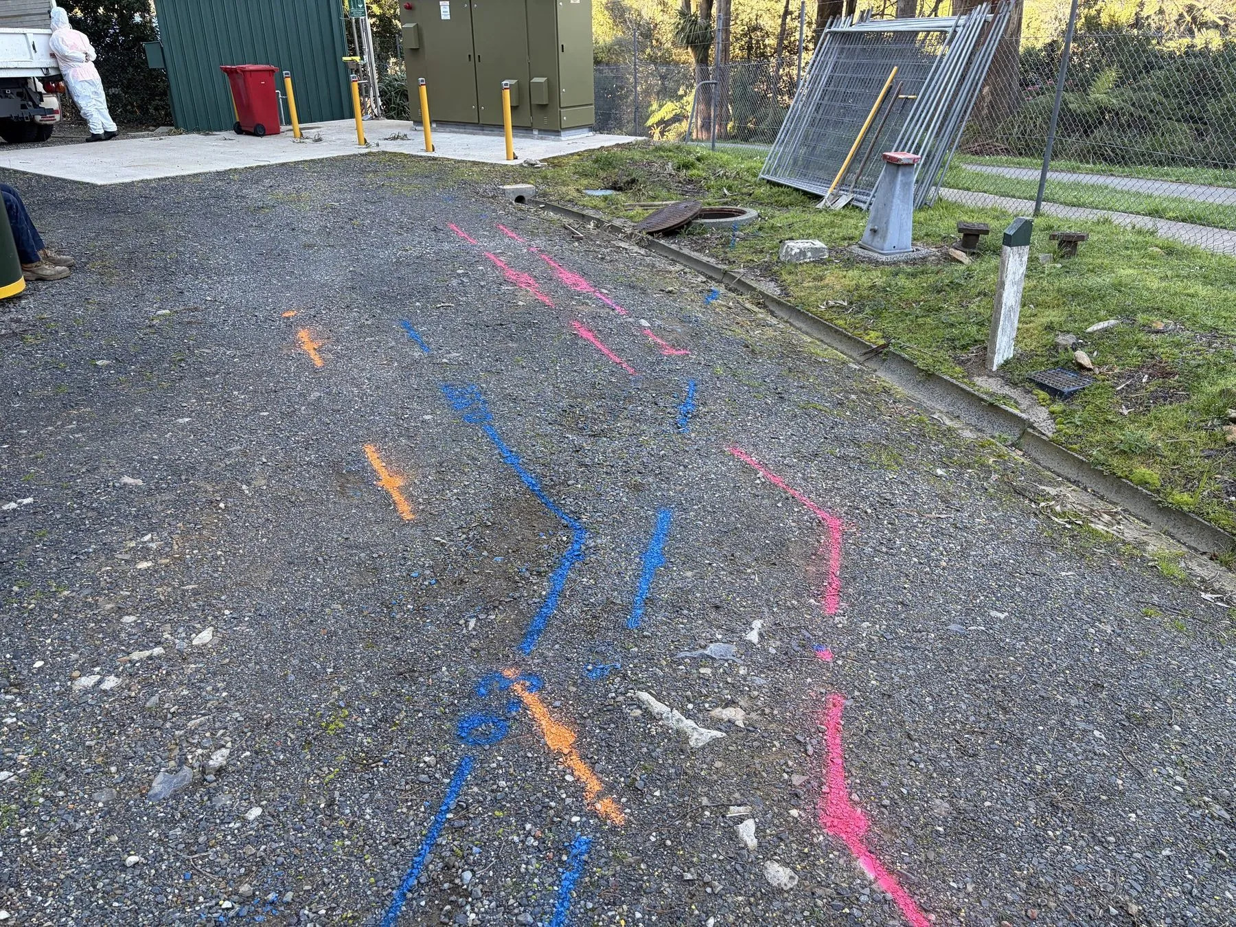 Blue, orange and pink locate lines sprayed across a gravel yard beside a grassy verge and a security fence