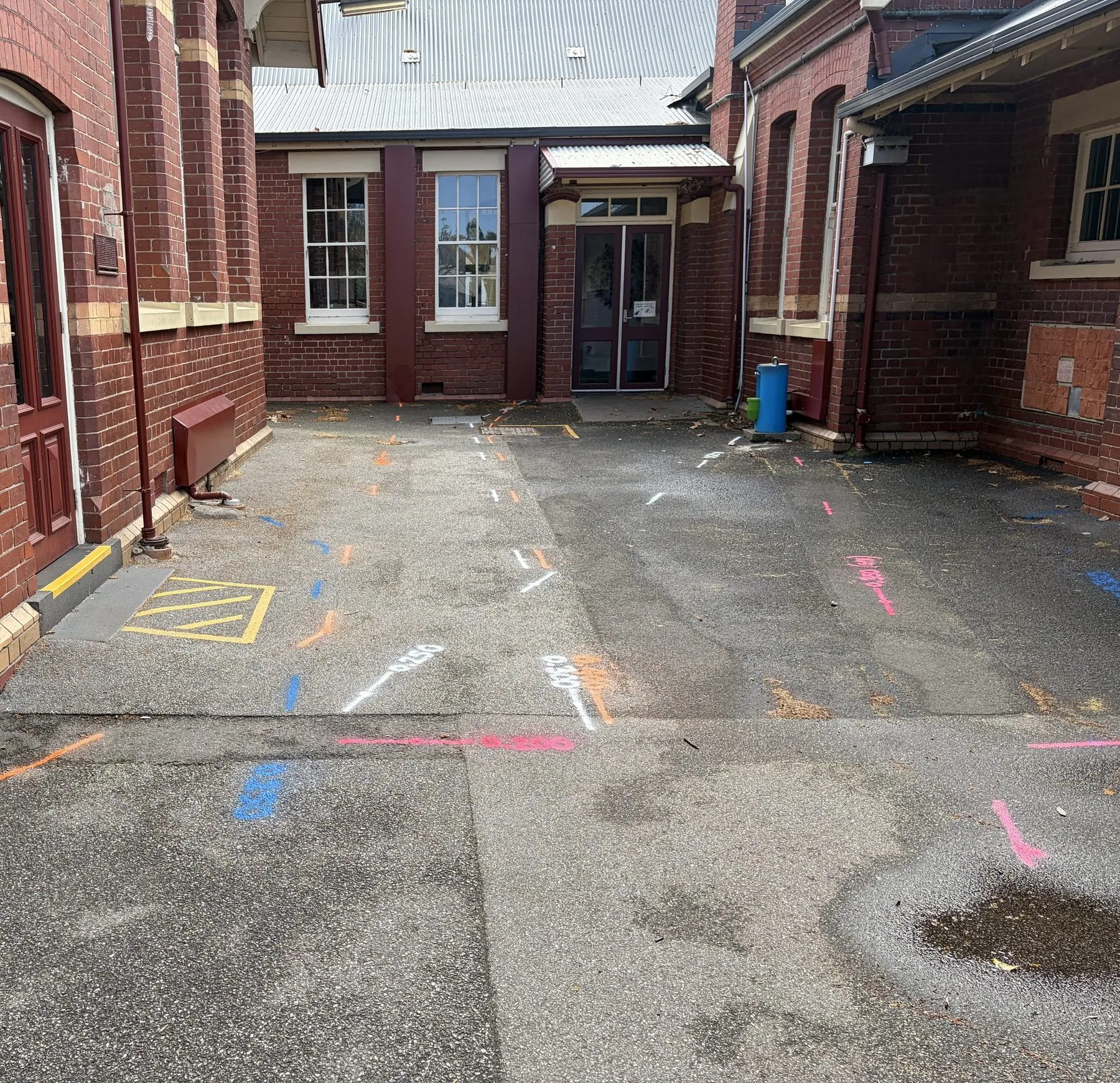 Blue, orange, white and pink locate marks with painted depths across an asphalt courtyard between red-brick buildings