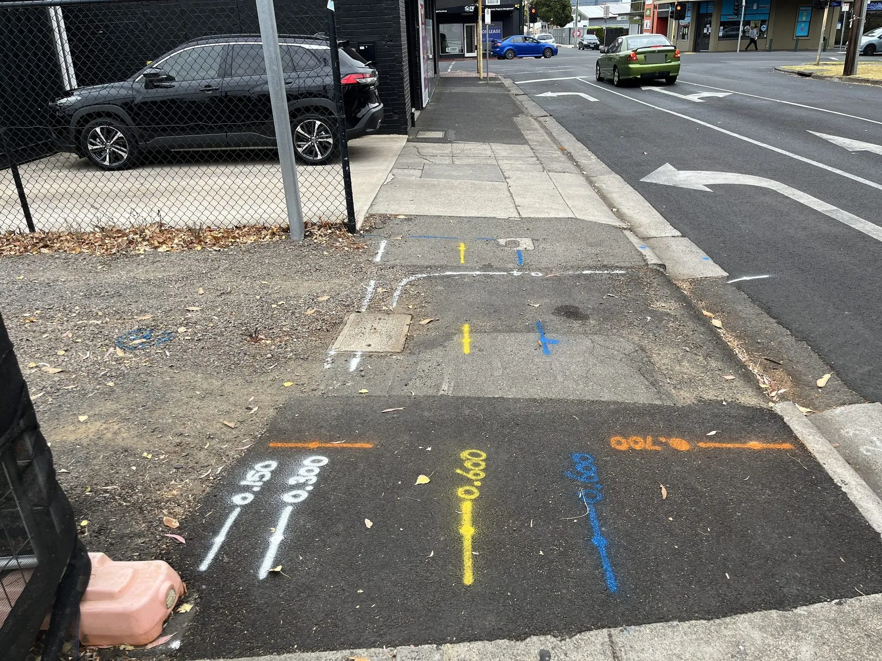 White, yellow, blue and orange locate lines with painted depths on fresh asphalt at the edge of a footpath beside a road
