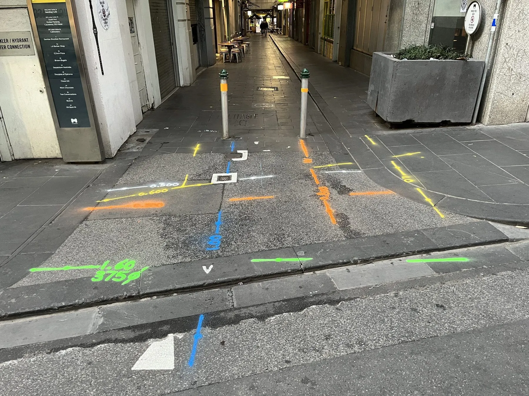 Yellow, orange, blue, green and white locate marks with painted depths sprayed across a city laneway entrance between two bollards
