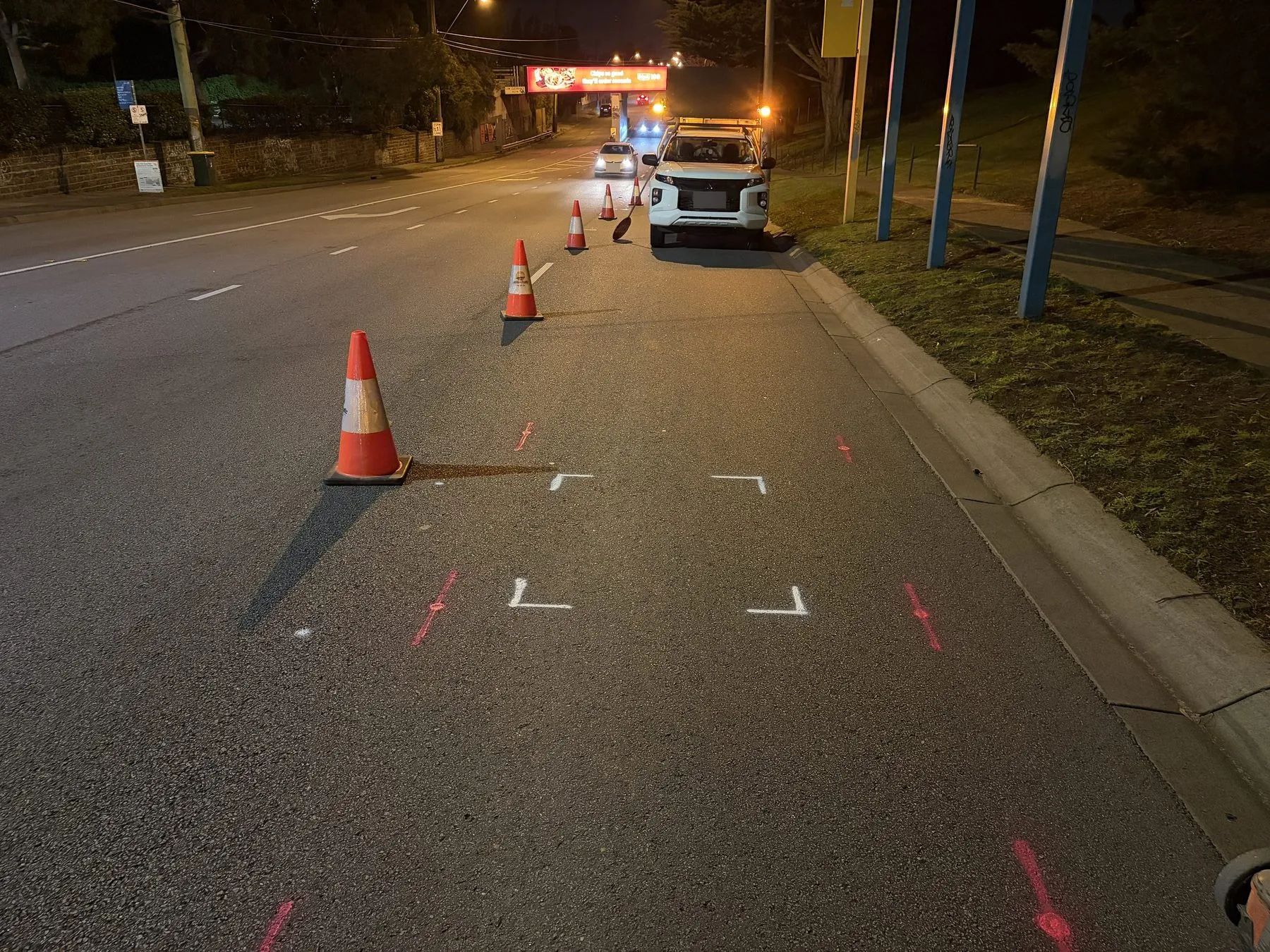 White corner marks outlining an area on a road at night, with red locate marks, traffic cones and a work ute