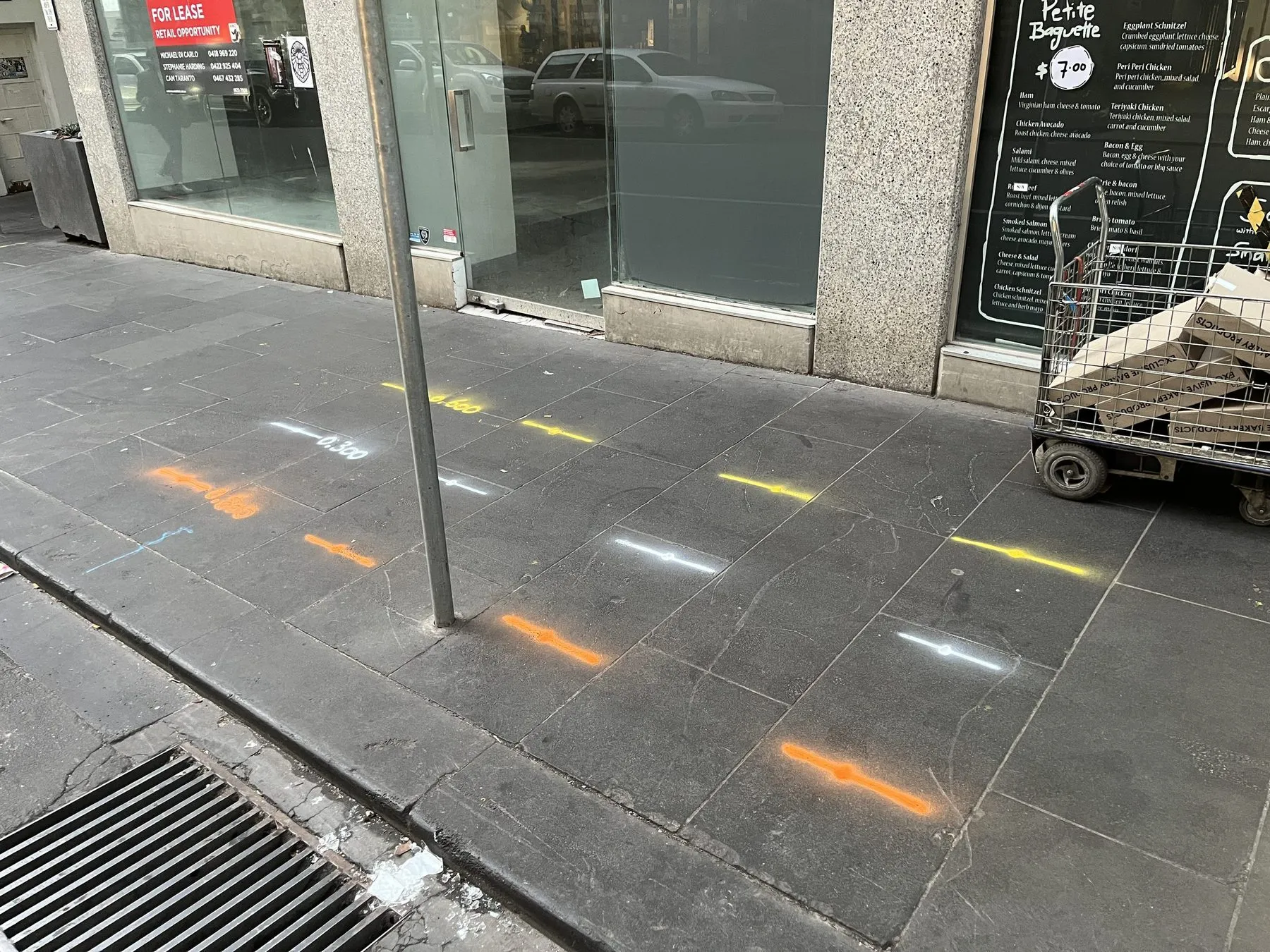 Orange, yellow and white locate marks with painted depths sprayed across the stone pavers of a city footpath