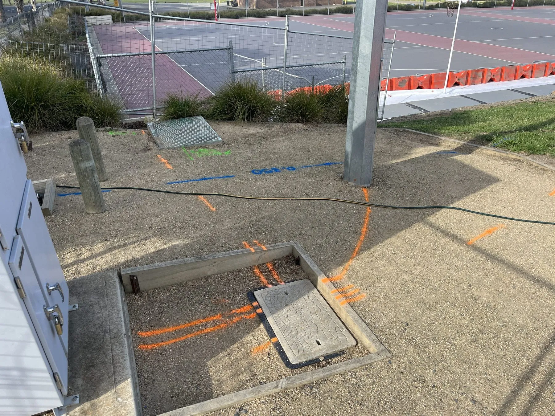 Orange locate lines traced into a pit beside an electrical cabinet, with a blue line and painted depth on the gravel behind