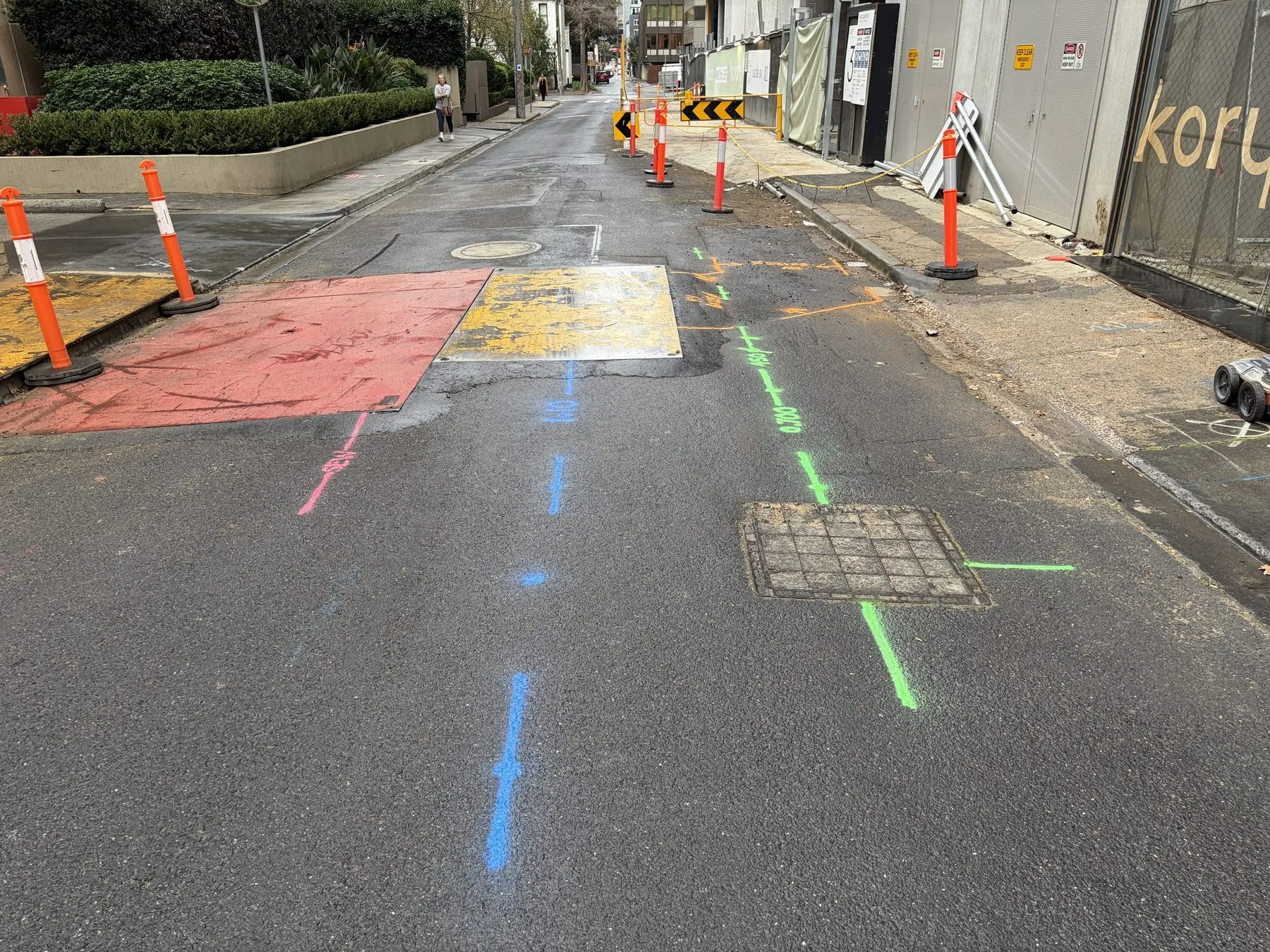 Blue, green and orange locate lines sprayed along a street works zone lined with traffic bollards, past a drainage pit